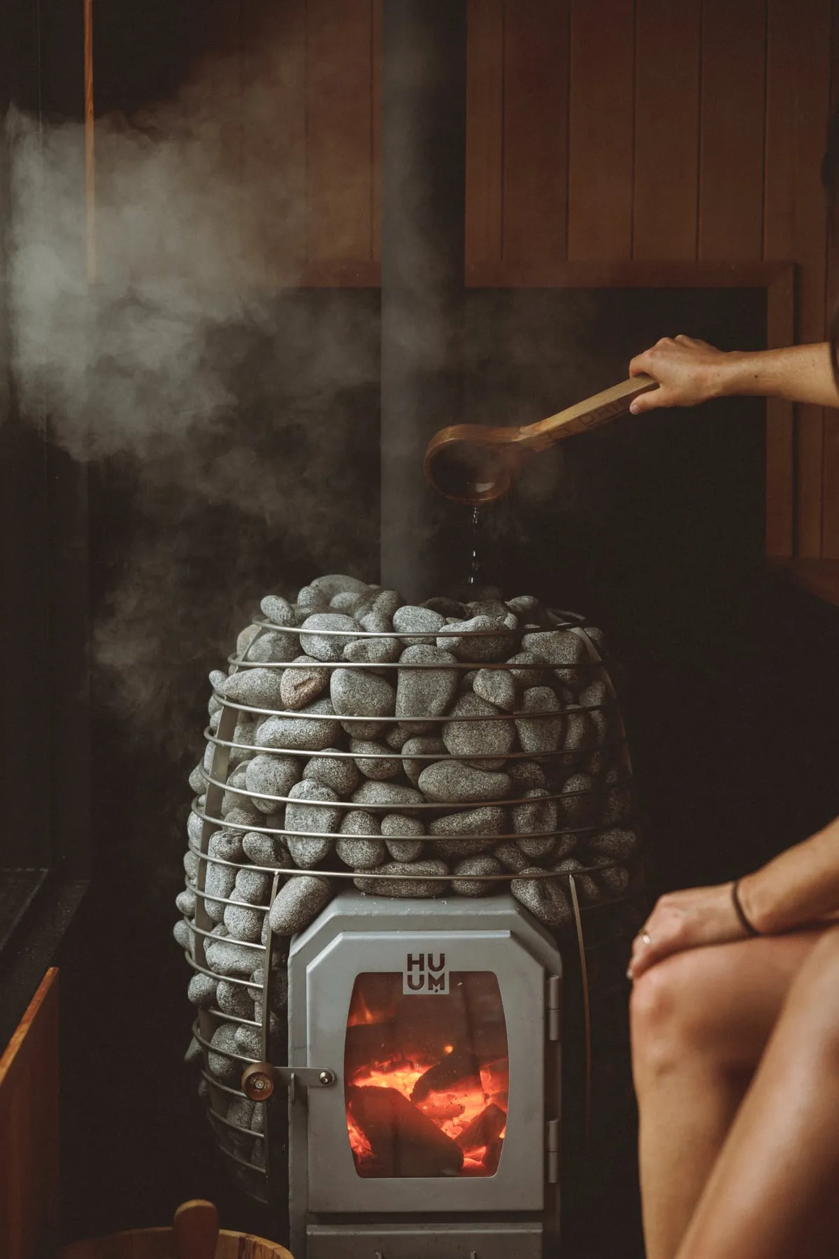 A ladle pouring water over the hot stones of a wood-fired sauna heater, steam rising